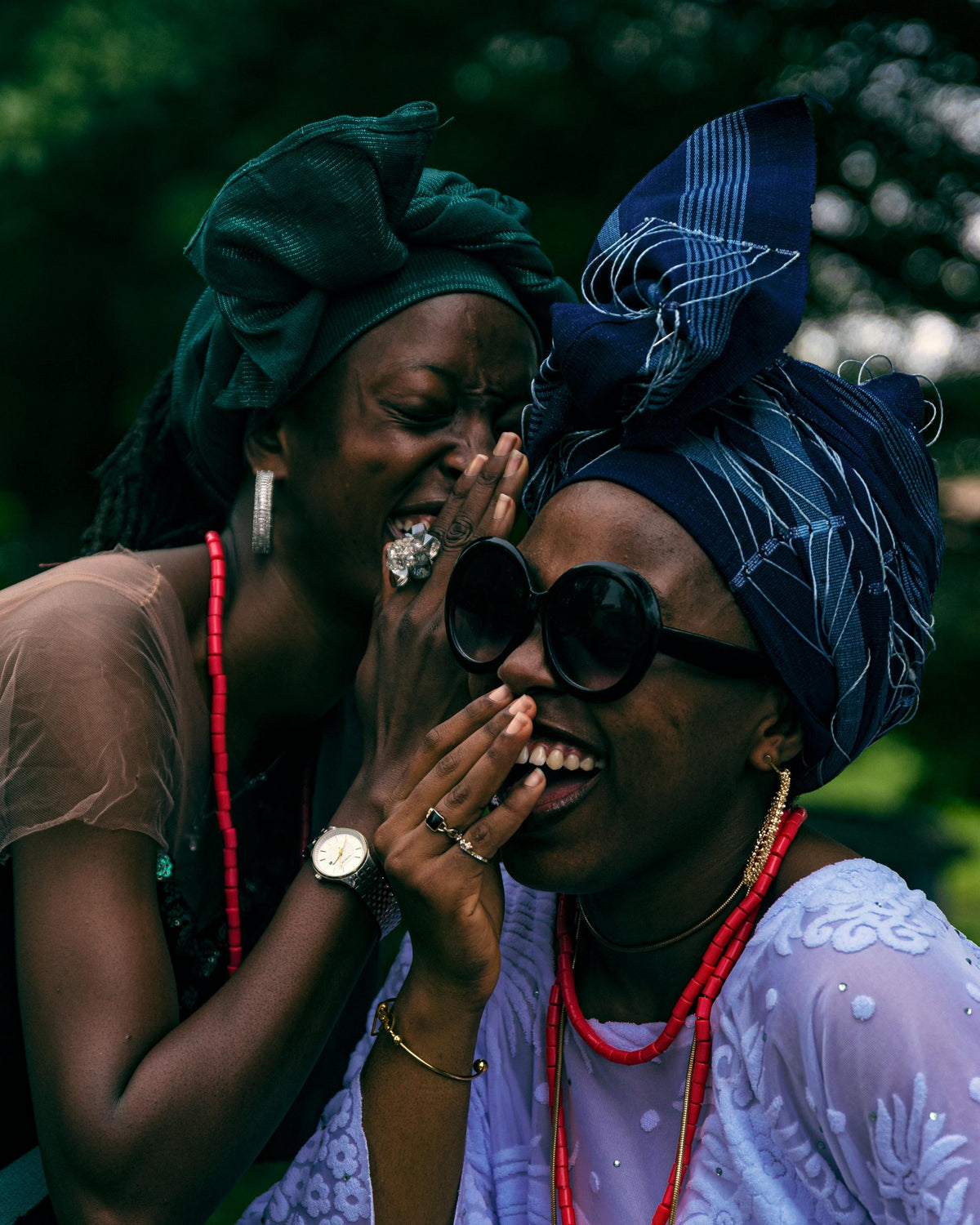Two women in head ties laughing together, one whispering to the other.