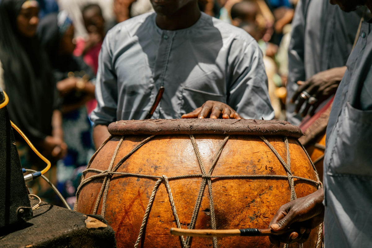 A man resting his hands on a large gourd drum with rope lacing, a crowd behind him.